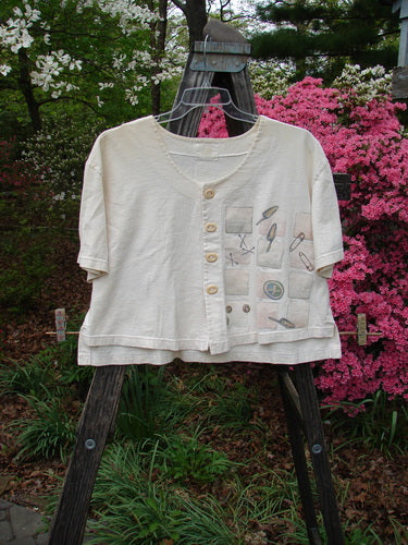 2001 Button Crop Top Pins and Needles Natural Size 1 displayed on a wooden easel, featuring unique buttons, wide short sleeves, and various pins, highlighting the garment's artistic and flowing design.