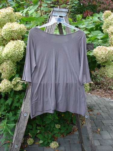 Barclay Batiste Raw Edge Layering Tee Unpainted Pebble Size 1, featuring three-quarter sleeves, a wide raw neckline, and rolled edges, hangs on a wooden A-frame, embodying Bluefishfinder's vintage aesthetic.