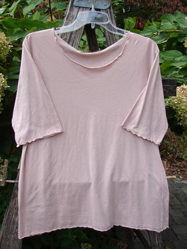 Barclay Batiste Curl Edge Layer Top Unpainted Blush Size 1, featuring a scooped neckline, scalloped hems, and almost three-quarter sleeves, hangs on a clear hanger amidst wooden supports and greenery.