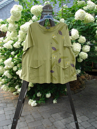 A green shirt with a design of birds on it, featuring a sweet ruffle hem and double drop pockets. The Barclay Double Pocket Twinkle Top Finch Leaf Peapod Size 2 is made from medium weight organic cotton.
