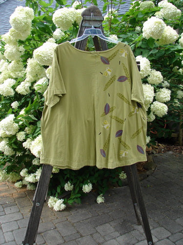 A green shirt with a design on it, featuring a sweet ruffle hem, banded lower sleeves, and double drop pockets. The shirt has an A-line shape and a softly rounded neckline. Made from medium weight organic cotton, this Barclay Double Pocket Twinkle Top in Peapod is in perfect condition. Size 2.