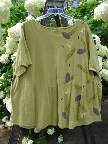 A Barclay Double Pocket Twinkle Top in Peapod, featuring a green shirt with a pattern on it. This medium weight organic cotton top has a sweet ruffle hem, banded lower sleeves, and double drop pockets. The A-line shape and softly rounded neckline add to its charm. Size 2.