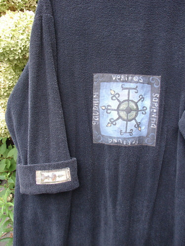 1999 Patched Chenille Angle Pocket Jacket Holiday Black Size 1, featuring a large embroidered compass patch on the back, matching sleeve patch, oversized button closure, and diagonal pockets against a shrubbery backdrop.