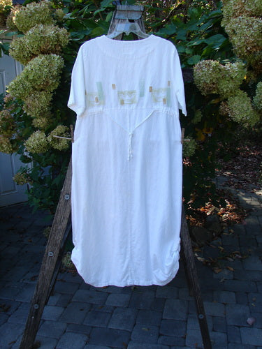 1999 Textured Linen Water Dress Tiny Fish White Size 0: A vintage-style white dress with pastel pocket panels, drawstring waist, and pleated front, hanging from a wooden ladder amid a garden setting.