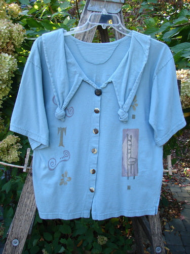 1994 Compass Top Chair Dusk Size 1: A light-blue, short-sleeved button-front top with a hood and decorative prints. Features include a pointed collar, original buttons, and a unique chair graphic motif.