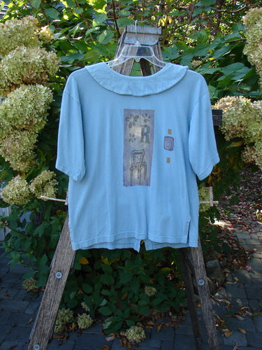1994 Compass Top Chair Dusk Size 1: Pale-blue women’s top with a Peter Pan collar, abstract graphic patch, and original BF buttons, hanging on a wooden ladder, reflecting Bluefishfinder.com's vintage style.