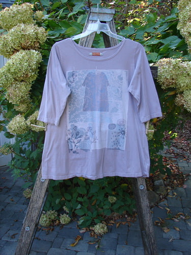 Barclay Three Quarter Sleeved Tee Play Dress Up Lilac Size 1, features floral and geometric motifs, displayed on a wooden ladder amidst greenery, highlighting its organic cotton fabric and unique neckline.