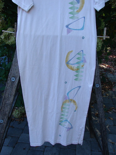 1996 Long Tea Dress Half Moon Birch Bark Size 0 displayed outdoors on a wooden frame, featuring pastel geometric motifs along one side. The dress has a rounded neckline, short wide sleeves, and a pegged vented hemline.