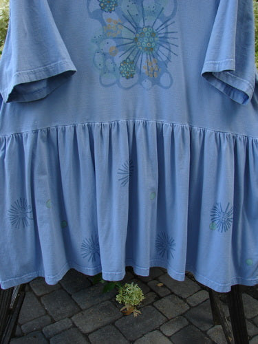 Barclay Collector's Tunic Dress Starburst Faded Sky Size 2: A blue shirt with a starburst pattern and dandelion flowers, featuring a drop waistline, exterior painted pockets, and a flouncy lower skirt.