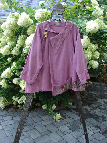 A close-up of a purple hemp jacket with a folded layered neckline and wooden toggle. The jacket has a varying hemline with horizontal pockets and sectional panels. It features a rear rounded collar, exterior stitchery, and a widening A-line shape. The lower sleeves are double stitched, and the jacket has a step stone paint design.