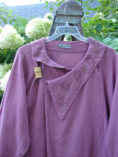 A purple hemp jacket with a folded layered neckline and a wooden toggle. Features include a fold-over front, varying hemline with pockets, and sectional panels.