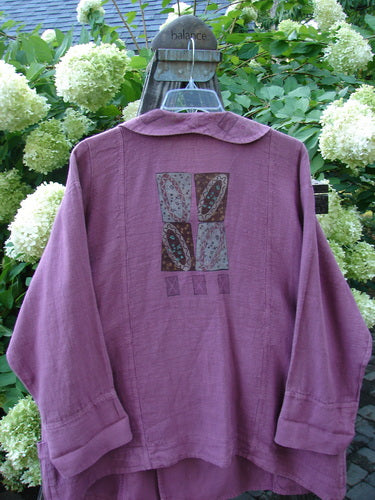 A purple hemp jacket with a folded layered neckline, oversized wooden toggle, and varying hemline with horizontal pockets. This substantial jacket features sectional panels, a rear rounded collar, exterior stitchery, and a widening A-line shape. The lower sleeves are double stitched, and it has suggestive step stone paint. Size 2.