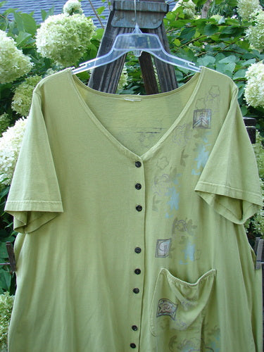 A Barclay Deep V Neck Button Pocket Cardigan in Sprout, featuring a mosaic floral theme paint. Made from medium-weight organic cotton, this size 2 cardigan has a deeply shaped neckline, wider short sleeves, and an A-line flair. The front hemline is varying and upward scooped, with an exterior drop side painted pocket. The cardigan also has alternating three sets of three button groupings. Bust: 56, Waist: 56, Hips: 60, Sweep: 75. Front Length: 33, Back Length: 37 inches.