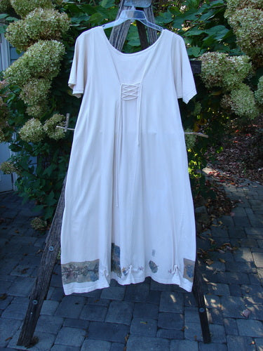 1994 Loop Dress Magic Garden Tea Dye Smaller OSFA displayed on a wooden ladder, showcasing its short sleeves, scoop neckline with lace-up detail, decorative appliqués, and flared hem against blooming hydrangeas.