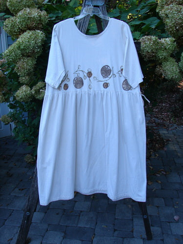 1994 Flower Garden Dress Garden Trail Altered Ecru Size 2: A knee-length white dress with a round neckline, embroidered midsection, gathered waist, and flowing lower section, hanging on a wooden stand outdoors.