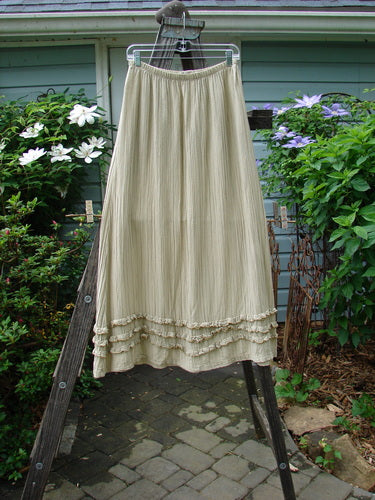 Barclay Rib Silk Triple Ruffle A Line Skirt in Sand, Size 0, displayed on a clothes rack outdoors with a plant in the background. Features a fluttery triple ruffle design and a silken texture. Vintage Blue Fish Clothing from BlueFishFinder.com.