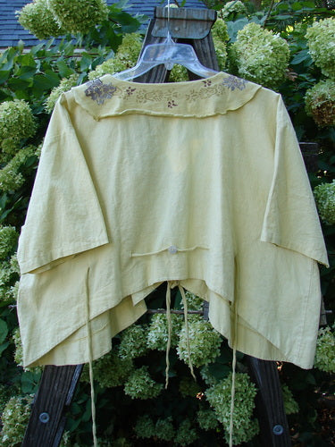 Image alt text: Barclay Linen Batiste Rippie Tie Jacket Shrug Florals Quiet Sunshine Size 2 - Yellow shirt with a swing silhouette, featuring a deep V-shaped neckline, longer front tie, painted flutter collar, rear tie back, varying hemline, and front pocket.