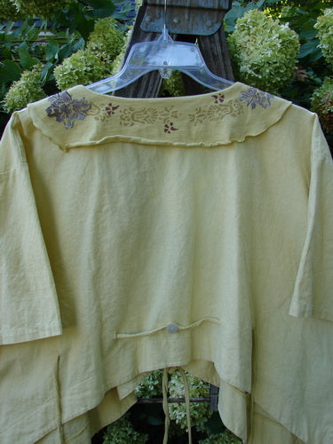 A yellow shirt with a floral design on it, featuring a deep V-shaped neckline, longer front batiste and cotton tie, painted batiste flutter collar, and a rippie rear tie back accented by a shell button. This Barclay Linen Batiste Rippie Tie Jacket Shrug in Quiet Sunshine is a sweet and stylish addition to your wardrobe.