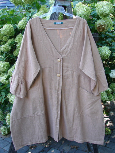 A beige Barclay Linen Adras Uptown Jacket in Pastel Rosewood, size 2, on a clothes rack. Features include a deep V-shaped neckline, sectional triangular panels, pleated and banded lower sleeves and hemline, double exterior drop front pockets, and a varying hemline. Unpainted for easy layering.
