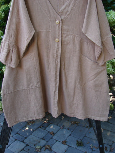A close-up of a Barclay Linen Adras Uptown Jacket in Pastel Rosewood, size 2. The jacket features a deep V-shaped neckline, sectional triangular panels, and pleated and banded lower sleeves and hemline. It has double exterior drop front pockets and a varying hemline with a rear skirted seam. Unpainted for easy layering.