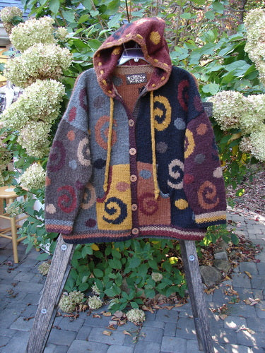 Tara Handknits Hooded Star Button Pocket Cardigan Swirls Brights OSFA, a handcrafted wool cardigan with patchwork and spiral motifs, hangs on a wooden ladder, showcasing its cozy design and vibrant details.