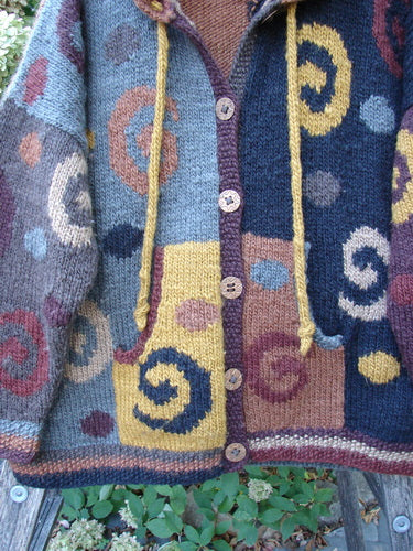 Tara Handknits Hooded Star Button Pocket Cardigan Swirls Brights OSFA draped over a wooden chair outdoors, showcasing its colorful patchwork, spiral motifs, oversized buttons, and drawstring hood.