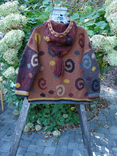 Tara Handknits Hooded Star Button Roo Pocket Cardigan Swirls Brights OSFA displayed outdoors; features intricate multicolored spiral patterns, ribbed accents, cozy funnel hood with tassel, and oversized metal star buttons on a wooden ladder.