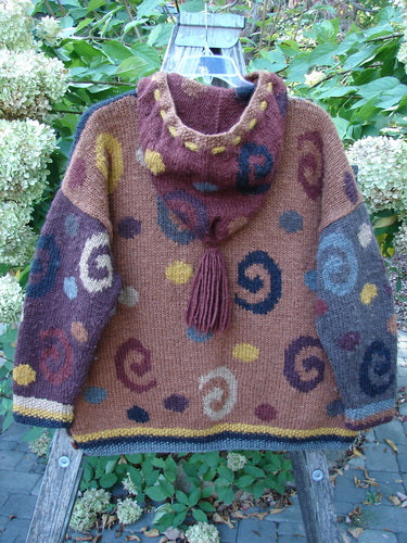 Tara Handknits Hooded Star Button Pocket Cardigan Swirls Brights OSFA displayed on a wooden stand in a garden. Features intricate spiral patterns, tassel hood, oversized star buttons, and kangaroo pocket.