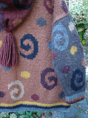 Tara Handknits Hooded Star Button Pocket Cardigan Swirls Brights OSFA features colorful spiral motifs and contrasting knit accents, with a cozy drawcord hood and oversized metal star buttons, hanging against foliage.
