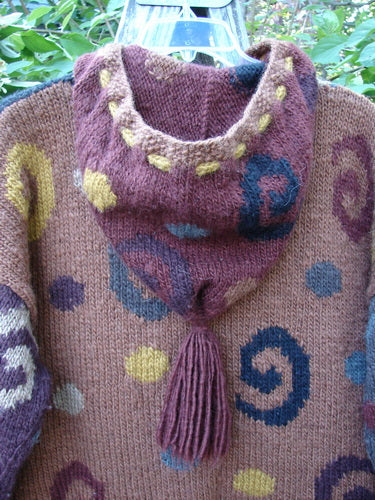 Tara Handknits Hooded Star Button Pocket Cardigan Swirls Brights OSFA features whimsical spiral motifs and oversized star buttons, hanging outdoors showcasing its vibrant handmade details and cozy hood with tassel.