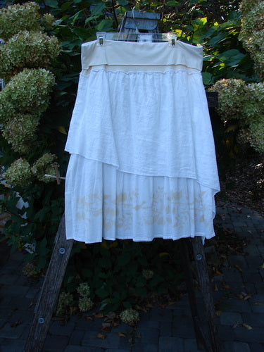 Barclay NWT Linen Fold Over Generosity Skirt on ladder, showcasing its tiered design and subtle vine and leaf detailing at the hem, against a garden backdrop of shrubs and hydrangeas.