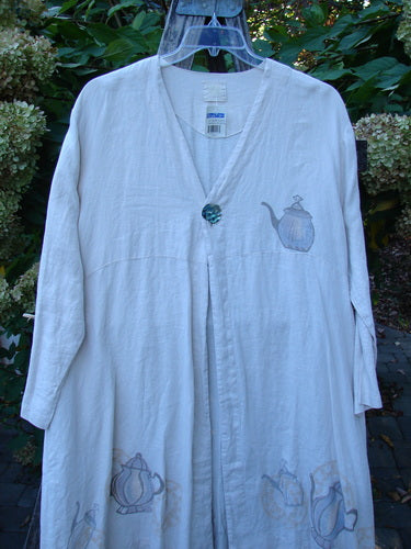1999 NWT Cream and Sugar Coat Tea Time Teadye OSFA: A medium-weight linen coat with a V-neck, decorative teapot motifs, oversized shell button, deep side pockets, and unique draping, hanging outdoors.