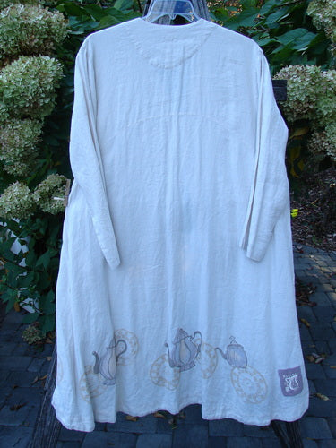1999 NWT Cream and Sugar Coat Tea Time Teadye OSFA: off-white linen tunic with back view, featuring teapot motif along hem, deep side pockets, and oversized shell button, hanging outdoors on a clear hanger.