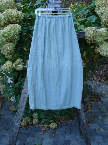 2000 Cross Dye Linen Contour Skirt Unpainted Meadow Size 0—ankle-length, pale grey-blue with elastic waist, slight A-line drape, and vertically paneled sides, hanging from a white hanger against a natural backdrop.