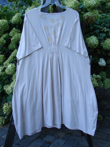 A Barclay Farmer Jen Tunic Dress in Pale Pink, featuring a white dress on a clothes rack. The dress has a flower theme paint in the lightest shade of pink, with generous measurements, oversized front drop pockets, and a deeper rounded neckline. Made from organic cotton, it is in perfect condition.