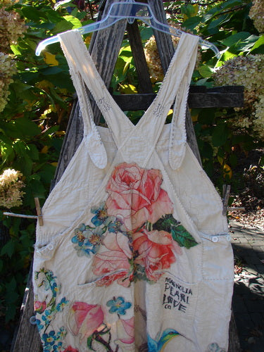 The Magnolia Pearl Applique Benjamin Overall Birdie Bouquet Moon OSFA features light-colored fabric adorned with pink roses and a bird motif, draped on a rustic ladder, highlighting its vintage charm.