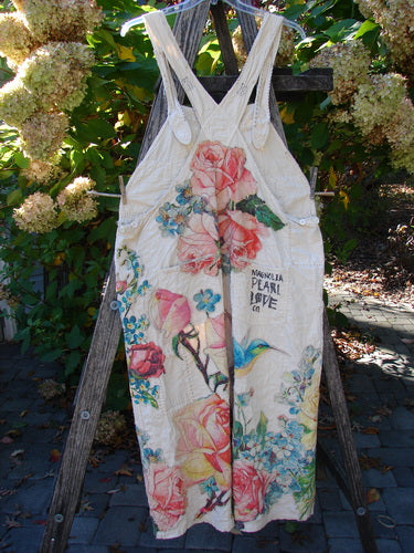 Magnolia Pearl Applique Benjamin Overall Birdie Bouquet Moon OSFA: Lightweight off-white dress with pink roses, blue flowers, and a hummingbird print, hanging on a rustic ladder, showcasing adjustable waist and multiple pockets.