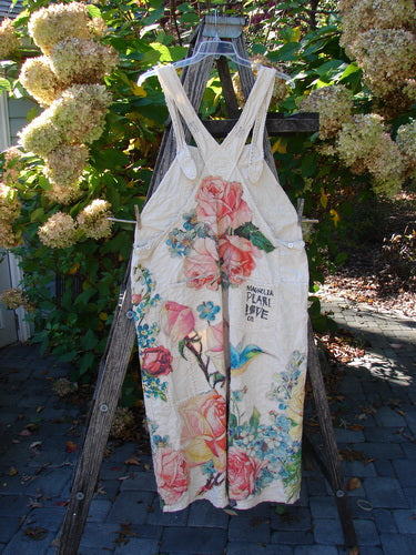 Magnolia Pearl Applique Benjamin Overall Birdie Bouquet Moon OSFA, a full-length sundress with wide straps, features a floral pattern, Plant Love text, and distinctive pockets, hanging on a ladder.