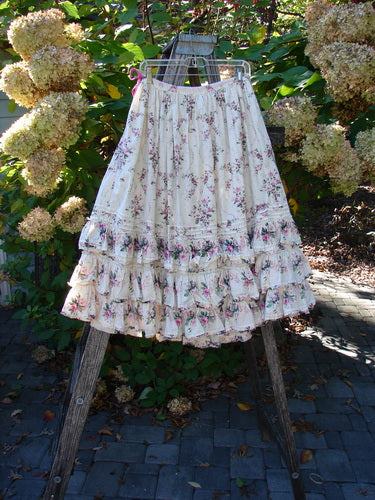 Magnolia Pearl Penelope Skirt Des Rosiers Floral Idealist OSFA, featuring a tiered design with floral print, ruffled hems, and lace accents, elegantly draped over a wooden ladder in a garden setting.
