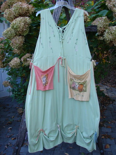1997 Salt Water Taffy Jumper Sea Life Mellon Size 2: Pastel-green, sleeveless dress with V-neck, drawstring ties, and two colorful pouch pockets with folk-art motifs, hanging outdoors against a hydrangea backdrop.