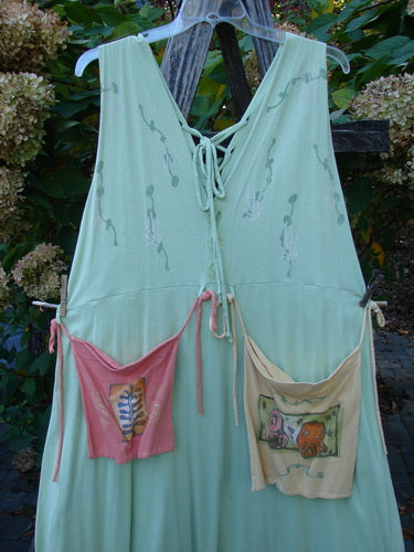 1997 Salt Water Taffy Jumper Sea Life Mellon Size 2: Light-green sundress with lace-up V-neck, subtle embroidery on bodice, and oversized patch pockets, featuring botanical and cartoon motifs, hanging against a garden backdrop.