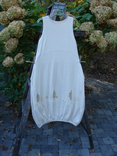 2000 Cotton Hemp Libby Jumper Falling Leaf Dove Altered Size 1, showcased on a wooden hanger outdoors, features subtle floral motifs at the hem, set against a backdrop of greenery and hydrangeas.