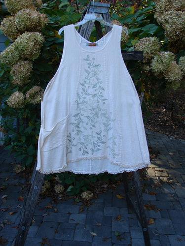 Barclay Slub Figure 8 Lace Pocket Pinafore Dress Soft Floral Crème Size 1 hanging on a wooden easel, featuring pale-green botanical accents, drop side pockets, and lace hemline, set against a garden backdrop.