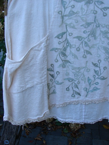 Barclay Slub Figure 8 Lace Pocket Pinafore Dress in Crème, featuring a stenciled vine-and-berry motif and lace hem. Displayed outdoors, highlighting its patch pocket and unique seams, perfect for expressing individuality.