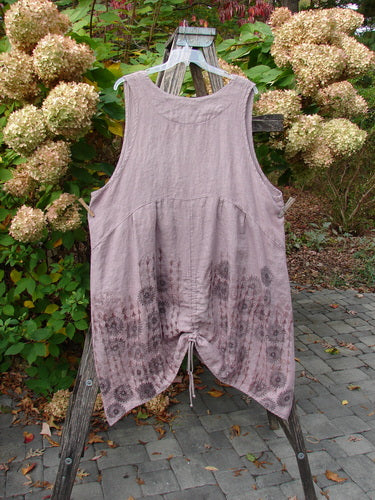 Barclay Linen Pleated Pocket Pinafore Mandala Soft Mauve Size 2, featuring an asymmetrical hem with dual drawcords and pleated empire waist, draped over a rustic ladder amidst garden blooms.