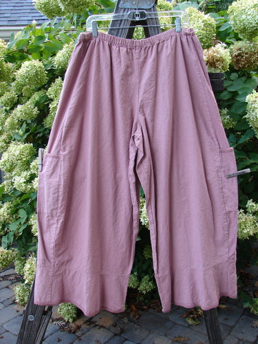 Image: A pair of pants on a clothes rack. The pants are from the Barclay Linen Spring Flutter Pant collection in Rosey Peony. They are size 2 and come unpainted. The pants have sectional panels, lower side drop pockets, and a larger elastic waistline. They feature sweet moving lower flutters, widening hips and leg, and a slight crop length. The waist measures 32 inches fully relaxed and 42 inches fully extended, while the hips measure 74 inches. The inseam is 27 inches and the length is 40 inches.
