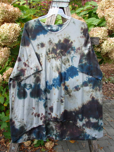 Barclay Drawcord Art Top Mottled Night Storm OSFA hangs on a clear plastic hanger, showcasing its loose fit, tie-dye pattern, rounded neckline, wide sleeves, and drawcord back amidst a garden setting.
