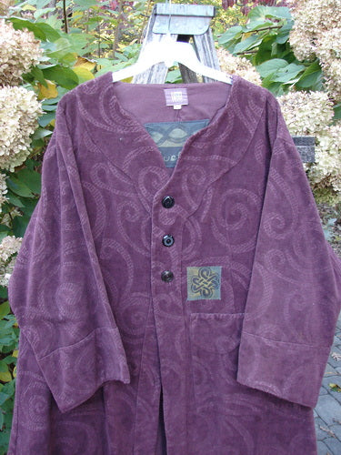 2000 Patched Upholstery Diwmach Coat Celtic Time Aubergine Size 2, featuring a swirl-pattern embossing, three dark buttons, decorative patch, scalloped neckline, and belled sleeves, displayed outdoors against lush garden foliage.