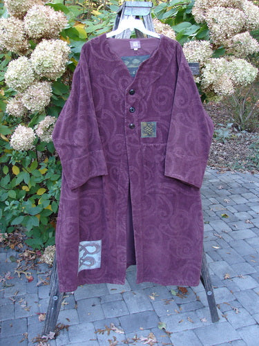 2000 Patched Upholstery Diwmach Coat Celtic Time Aubergine Size 2 displayed outdoors, showcasing its loose duster style, damask swirl patterns, and decorative Celtic appliqué patches against a rustic garden setting.