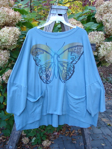 Barclay NWT Two Pocket Mariposa Pullover Butterfly Sea OSFA features an oversized butterfly graphic, drop-shoulder sleeves, and front pockets, hanging on a ladder in a garden setting.